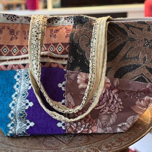 Peut inclure: Un sac fourre-tout patchwork avec divers tissus à motifs dans des tons de marron, bleu, violet et imprimés floraux. Le sac a des anses beiges et est posé sur un plateau en métal décoratif.