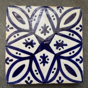 May include: A white ceramic tile with a blue and white geometric pattern. The pattern features a central star shape surrounded by four stylized flower shapes.