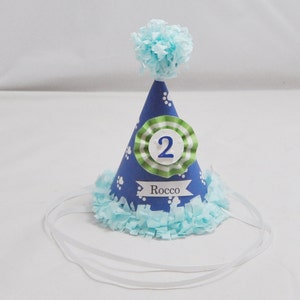 May include: Blue and green party hat with white paw prints and a white ribbon. The hat has a white circle with the number "2" in green and the name "Rocco" written below. A blue pom-pom sits on top of the hat.