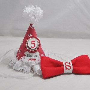 May include: Red and white dog party hat with a white pom pom on top. The hat has white paw prints and a white bone with the name "Sinatra" on it. The hat also has a white and red ribbon with the number "5" on it. A red bow tie with a white "S" on it is also included.