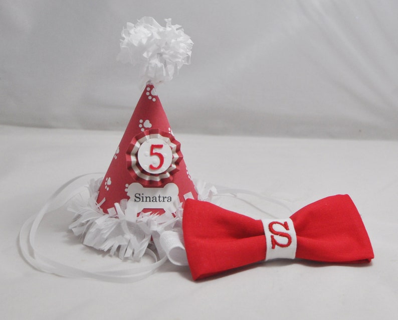 May include: Red and white dog party hat with a white pom pom on top. The hat has white paw prints and a white bone with the name "Sinatra" on it. The hat also has a white and red ribbon with the number "5" on it. A red bow tie with a white "S" on it is also included.