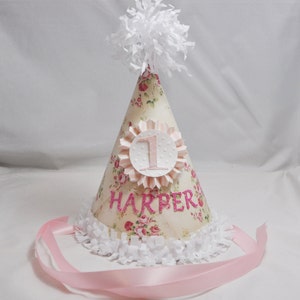 May include: A pink and white party hat with a floral pattern and the name "HARPER" and the number "1" on it. The hat has a white pom-pom on top and a pink ribbon.