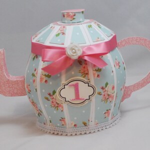 May include: A blue and pink teapot with a floral pattern and a pink ribbon. The teapot has a white lace trim and a number "1" on the front.