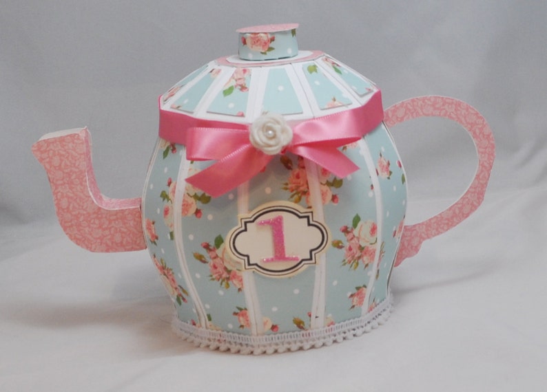 May include: A blue and pink teapot with a floral pattern and a pink ribbon. The teapot has a white lace trim and a number "1" on the front.