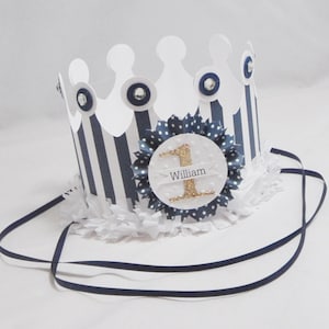 May include: A white paper crown with blue and white stripes, a white and blue polka dot circle with a gold glitter number one and the name "William" on it. The crown has a white ruffle trim and a black ribbon.