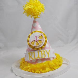 May include: Pink and white polka dot party hat with a yellow lemon slice and the number 1 on it. The hat has a yellow tissue paper pom pom on top and a white ribbon around the bottom. The hat says "RUBY" on the front.