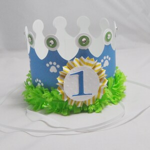 May include: A blue and white paper crown with paw prints and a yellow and white striped bow. The crown has a white circle with the number "1" in blue on it. The crown is on a white background.