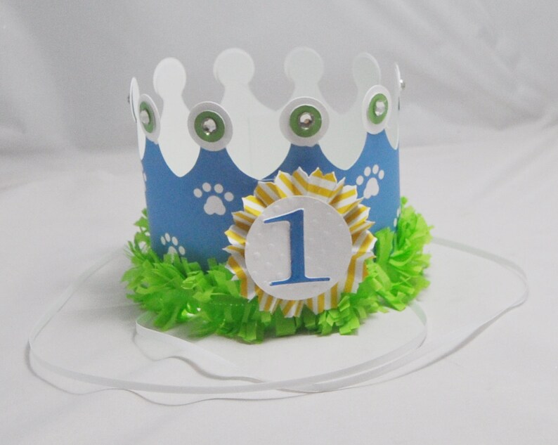May include: A blue and white paper crown with paw prints and a yellow and white striped bow. The crown has a white circle with the number "1" in blue on it. The crown is on a white background.