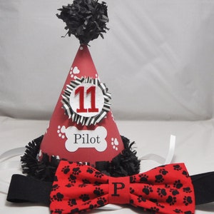 May include: Red and black birthday party hat with a black pom-pom on top. The hat has a white circle with the number 11 in red and a bone with the word "Pilot" on it. A red bow tie with black paw prints is included.