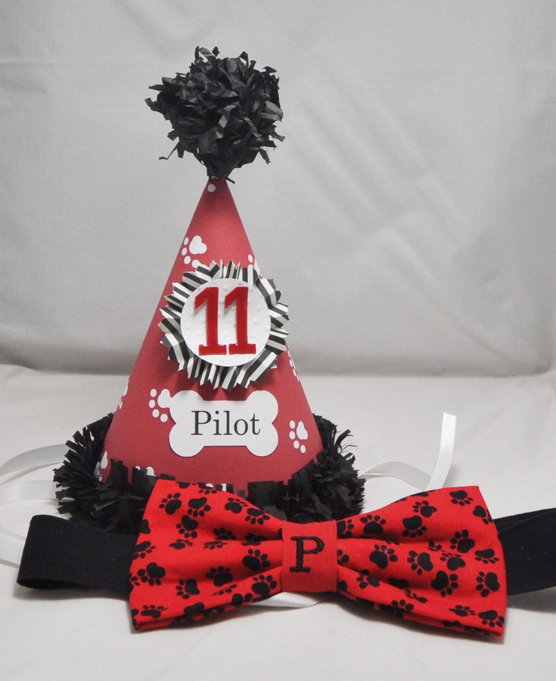 May include: Red and black birthday party hat with a black pom-pom on top. The hat has a white circle with the number 11 in red and a bone with the word "Pilot" on it. A red bow tie with black paw prints is included.