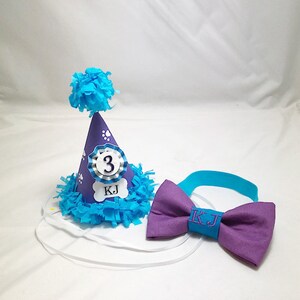May include: Purple and blue dog birthday party hat with a white bone and the text "KJ" and the number "3". A matching purple bow tie with the text "KJ" in turquoise.