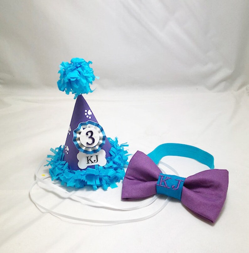 May include: Purple and blue dog birthday party hat with a white bone and the text "KJ" and the number "3". A matching purple bow tie with the text "KJ" in turquoise.
