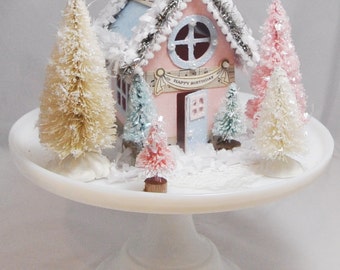 May include: A pink and blue miniature house with a white star on top, surrounded by miniature white, pink, and blue trees. The house has a sign that says "Happy Birthday". The house is sitting on a white cake stand.
