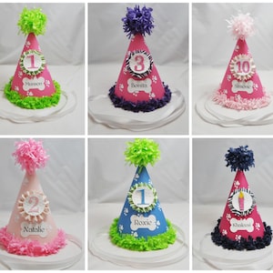 May include: Six pink, blue, and white dog birthday hats with paw print designs and numbers 1, 2, 3, and 10. Each hat has a name on it: Munson, Bonita, Simone, Natalie, Roxie, and Khaleesi.