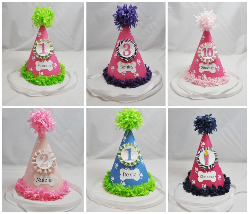 May include: Six pink, blue, and white dog birthday hats with paw print designs and numbers 1, 2, 3, and 10. Each hat has a name on it: Munson, Bonita, Simone, Natalie, Roxie, and Khaleesi.