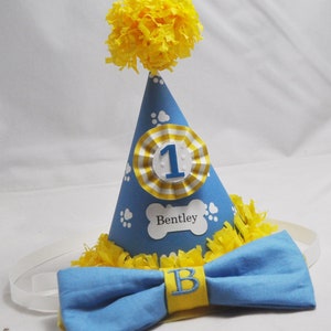 May include: A blue and yellow dog-themed party hat with a white paw print design and the number "1" on the front. The hat has a yellow pom-pom on top and a matching blue bow tie with a yellow "B" embroidered on it.