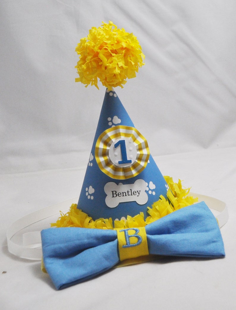 May include: A blue and yellow dog-themed party hat with a white paw print design and the number "1" on the front. The hat has a yellow pom-pom on top and a matching blue bow tie with a yellow "B" embroidered on it.