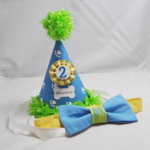 May include: A blue and yellow dog birthday party hat with a white paw print design and the number "2" on the front. The hat has a green pom-pom on top and the name "Maximus" is written on the front. A blue bow tie with a green and yellow stripe and the letter "M" embroidered on it is included.