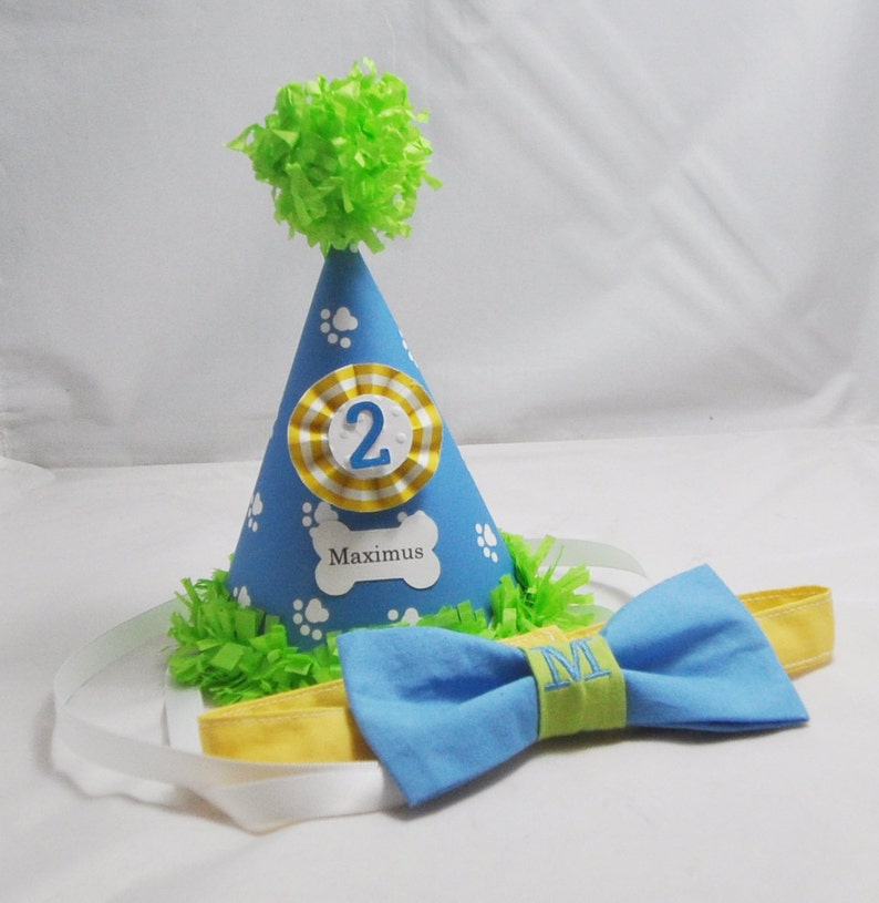 May include: A blue and yellow dog birthday party hat with a white paw print design and the number "2" on the front. The hat has a green pom-pom on top and the name "Maximus" is written on the front. A blue bow tie with a green and yellow stripe and the letter "M" embroidered on it is included.