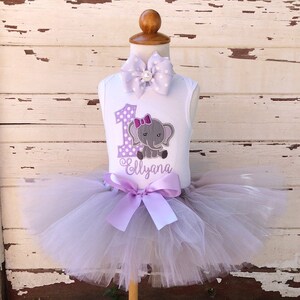 May include: A white sleeveless onesie with a purple polka dot number one and a gray elephant with a purple bow. The onesie has the name "Ellyana" embroidered on it. The onesie is paired with a lavender tulle tutu.