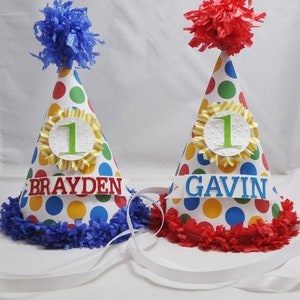 以下が含まれることがあります： 白い水玉模様、赤と青のフリンジ、そして「Brayden」と「Gavin」という名前が書かれた、カラフルな誕生日パーティーハット2つ。それぞれのハットには、黄色と白のストライプの円の中に緑色の数字「1」が書かれています。