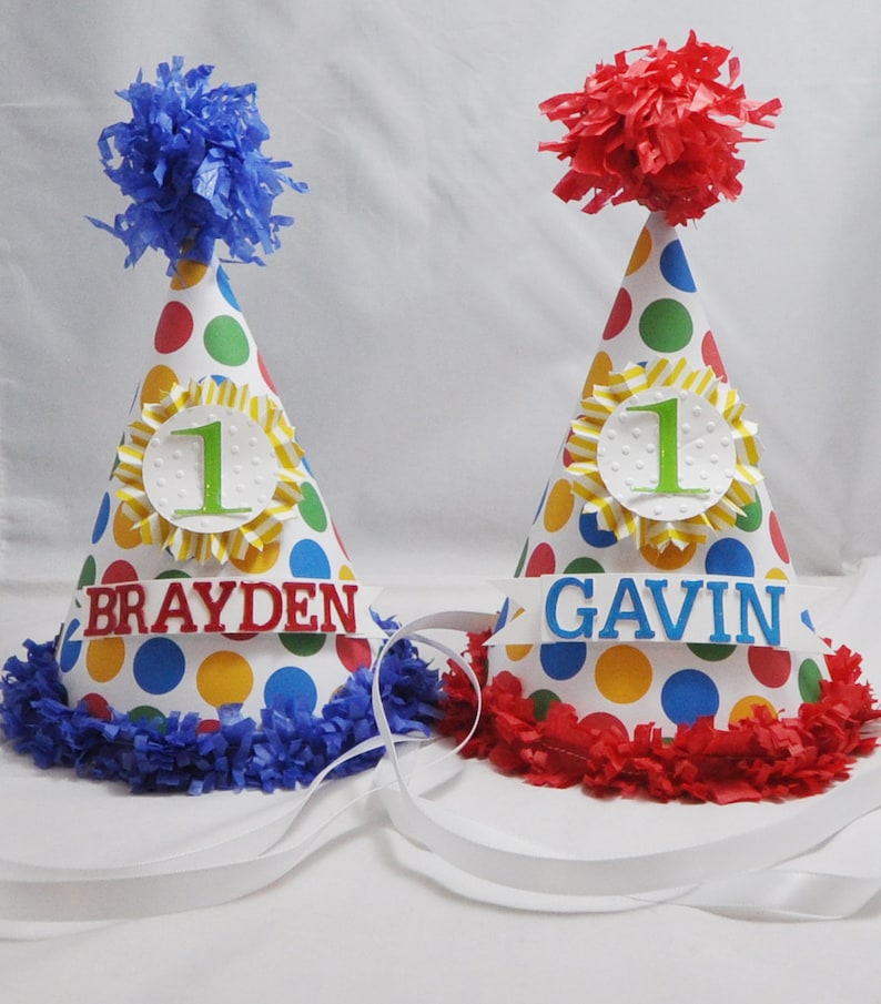 以下が含まれることがあります： 白い水玉模様、赤と青のフリンジ、そして「Brayden」と「Gavin」という名前が書かれた、カラフルな誕生日パーティーハット2つ。それぞれのハットには、黄色と白のストライプの円の中に緑色の数字「1」が書かれています。