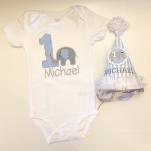 May include: A white onesie with a blue number one and a gray elephant. The onesie is personalized with the name "Michael". A blue and white striped party hat with a gray elephant and the number one is also personalized with the name "Michael".