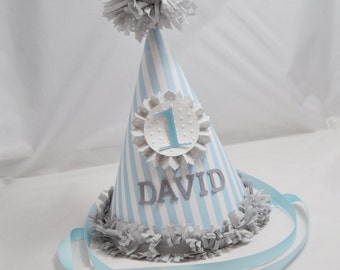 May include: A blue and white striped party hat with a gray pom-pom on top. The hat has a white circle with gray polka dots and the number "1" in the center. The name "DAVID" is written in gray letters on the front of the hat.