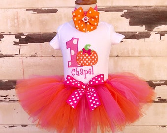 May include: A white short-sleeved bodysuit featuring a pink number one, an orange polka-dot pumpkin, and the name Chapel in pink embroidery. A multi-layered orange and pink tulle tutu with a large pink polka-dot bow and a matching hair bow.