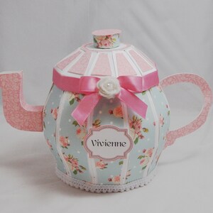 May include: A light blue and pink teapot with a white lace trim, a pink ribbon, and a white flower. The teapot has a floral pattern and the name "Vivienne" is written on a label.