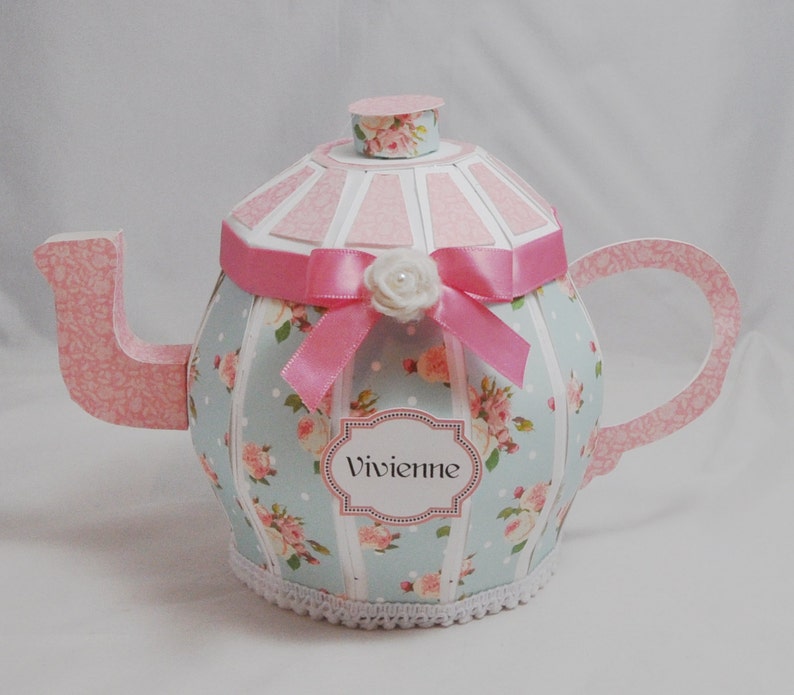 May include: A light blue and pink teapot with a white lace trim, a pink ribbon, and a white flower. The teapot has a floral pattern and the name "Vivienne" is written on a label.
