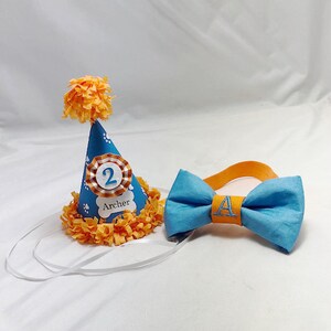 May include: A blue and orange birthday party hat with a white paw print design and the number "2" and the name "Archer" printed on it. A blue bow tie with an orange center and the letter "A" embroidered on it.