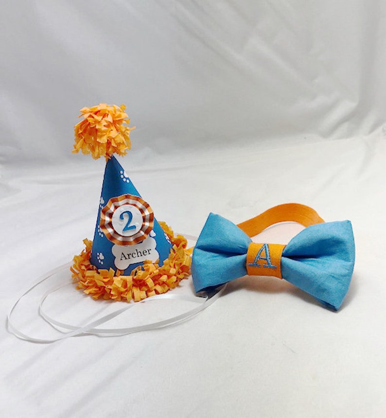 May include: A blue and orange birthday party hat with a white paw print design and the number "2" and the name "Archer" printed on it. A blue bow tie with an orange center and the letter "A" embroidered on it.