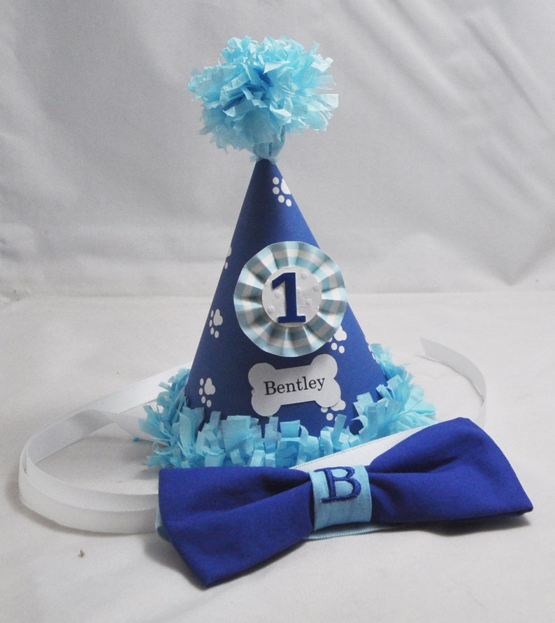 May include: Blue and white dog birthday party hat with a white bone-shaped tag that says "Bentley" and a blue bow tie with a white "B" on it.