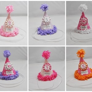 May include: Set of ten pink, purple, and orange dog party hats with white paw print designs. Each hat has a white ribbon, a white pom-pom, and a white circle with a number and a name. The names are Ellie, Paris, Bella, Lucy, Posey, Pumpkin.