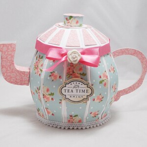 May include: A blue and pink paper teapot with a white lace trim, decorated with pink roses and white polka dots. The teapot has a pink ribbon with a white flower and a label that says "Steep stir Tea Time Enjoy".