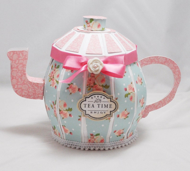 May include: A blue and pink paper teapot with a white lace trim, decorated with pink roses and white polka dots. The teapot has a pink ribbon with a white flower and a label that says "Steep stir Tea Time Enjoy".