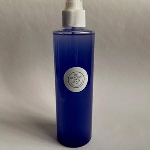 Pode incluir: Um frasco de spray azul com uma bomba branca e uma tampa transparente. O frasco contém um líquido azul e tem um rótulo branco com texto. O produto é provavelmente um artigo cosmético ou de aromaterapia.