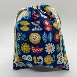 May include: A drawstring bag with a vibrant floral pattern in yellow, red, white, and blue. The bag features a navy blue background with stylized flowers, leaves, and butterfly designs. The drawstring is a light gold color.
