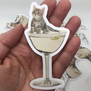 May include: A sticker of a kitten sitting in a martini glass. The glass is filled with a clear liquid and a lemon wedge. The sticker is white with a black outline and the artist's signature, "M. Shields", is at the bottom.