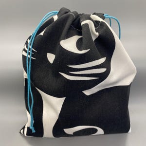 May include: A black and white drawstring bag featuring a cat design. The bag is made of fabric and has a repeating pattern of a cat's face and body. The drawstring is a bright blue color.