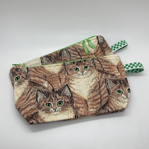 May include: Two rectangular fabric pouches with a repeating pattern of brown tabby cats with green eyes. The pouches have green zippers and green and white checkered ribbon loops. The background is white.