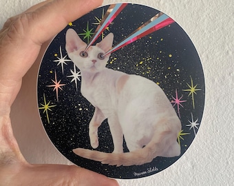 Op de afbeelding: Een ronde sticker met een witte kat met roze accenten, tegen een zwarte achtergrond met gele en witte stippen. De kat heeft kleurrijke stralen die uit zijn ogen komen, met stervormige accenten. De handtekening van de kunstenaar staat rechtsonder.