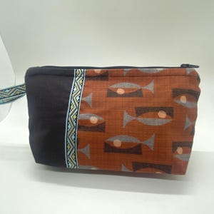 May include: A rectangular pouch with a black zipper and a patterned design. The pouch features a brown fabric with a fish pattern and a black fabric on the left side. A decorative trim and a small strap are also visible.