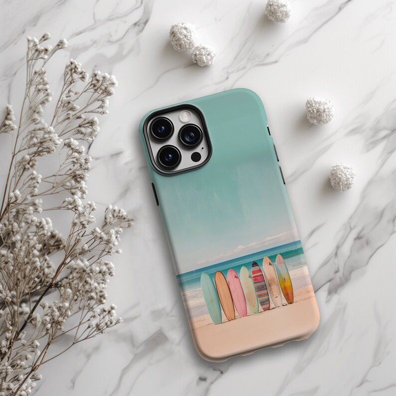 An iPhone case with surfboards standing on a beach by the ocean lays on a white marble background with 5 white cottony cluster balls in the upper right hand corner. On the left side of the image are springs of white wildflowers