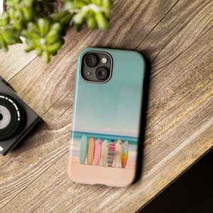 An iPhone case with surfboards standing on a beach by the ocean lays on a wood table, top view, with a portion of a green plant in the upper left hand corner and a portion of a black old school camera is on the left hand side of the image