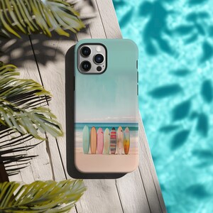 An iPhone case with surfboards standing on a beach by the ocean lays on a light washed wood table above a pool with a green palm plant on the bottom lefthand side