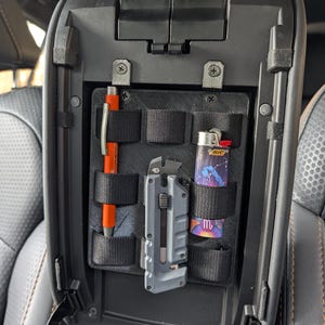 Pode incluir: Organizador de consola de carro preto com vários compartimentos. Inclui uma caneta laranja, um canivete e um isqueiro com um design de constelação. O organizador foi projetado para guardar itens pequenos com segurança.