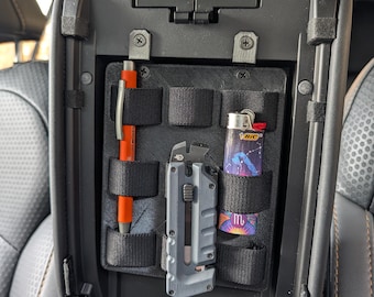 May include: Black car console organizer with multiple compartments. Includes an orange pen, a utility knife, and a lighter with a constellation design. The organizer is designed to hold small items securely.