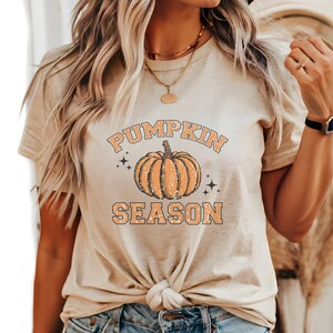 May include: A beige short-sleeve t-shirt featuring a graphic of an orange pumpkin in the center. The text PUMPKIN SEASON is printed in orange block letters above and below the pumpkin, accented by small black star icons on both sides.